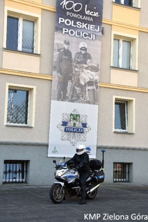 zdjęcie kolorowe, policjant w białym kasku motocyklowym siedzący na policyjnym motocyklu na tle banneru z napisem &quot;100-lecie powołania Polskiej Policji&quot;