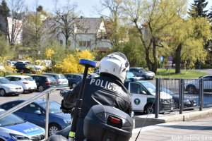 zdjęcie kolorowe, policjant w białym kasku motocyklowym siedzący na policyjnym motocyklu na tle samochodów stojących na parkingu. Na plecach policjanta widoczny napis &quot;policja&quot;.