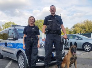 Dwóch policjantów wraz z psem policyjnym pozujących do zdjęcia na tle radiowozu