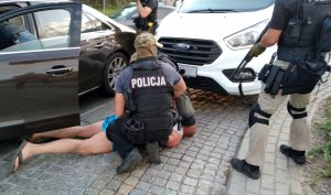Zamaskowany policjant zakuwa w kajdanki mężczyznę leżącego na chodniku pomiędzy autami