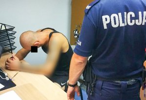 Policjant stoi i pilnuje zatrzymanego który siedzi i chowa głowę przed zdjęciem