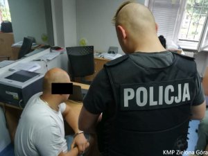 Policjant stoi nad siedzącym mężczyzną w kajdankach przy biurku