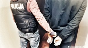 na zdjęciu widoczny jest policjant po cywilnemu w czarnej kamizelce  z napisem Policja. obok niego inny mężczyzna - widoczne dłonie z kajdanakami założonymi na ręce trzymane z tyłu. Policjant prowadzi mężczyznę trzymając za kajdanki