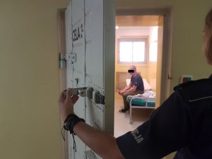 Funkcjonariuszka policji otwierająca drzwi do celi, w której na skraju łóżka siedzi mężczyzna.