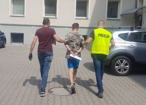 W słoneczny dzień, dwóch funkcjonariuszy policji prowadzi, po parkingu policyjnym, młodego mężczyznę ubranego w koszulkę moro i niebieskie krótkie spodenki.