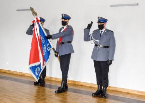 Trzech policjantów stoi na tle białej ściany, gdzie środkowy trzyma sztandar Rzeczypospolitej Polski a pozostali salutują.