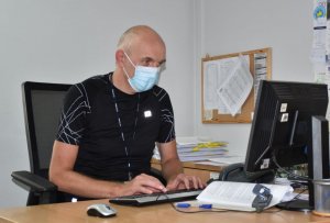 Starszy funkcjonariusz policji, z maseczką ochronną zakrywającą usta oraz nos, pisze na czarnej klawiaturze komputera stacjonarnego.