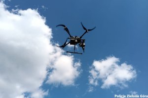 czarny dron lecący na tle błękitnego nieba z białymi obłokami