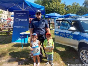 Policjantka z dwójką dzieci stoi na tle radiowozu i stoiska komendy miejskiej policji